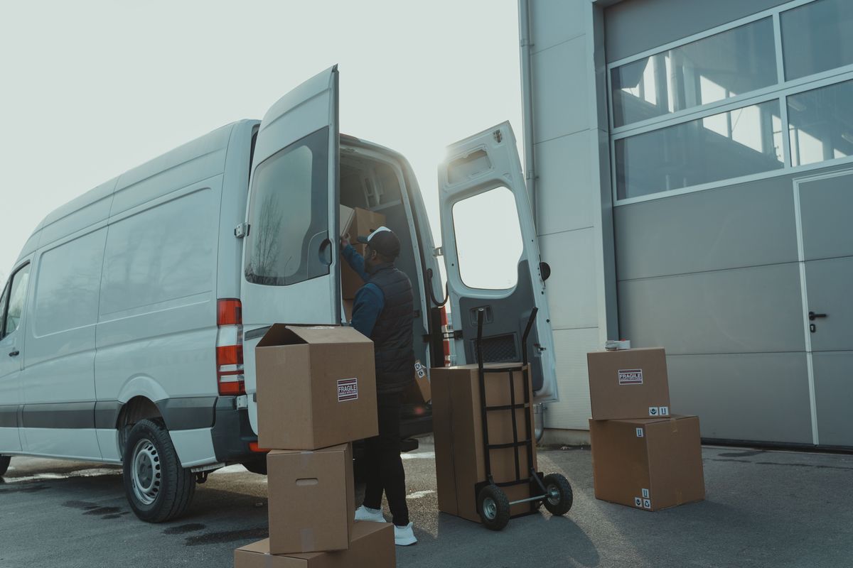 Een koerier laadt dozen in een witte bestelwagen met open achterdeuren, naast een steekkar met een grote doos